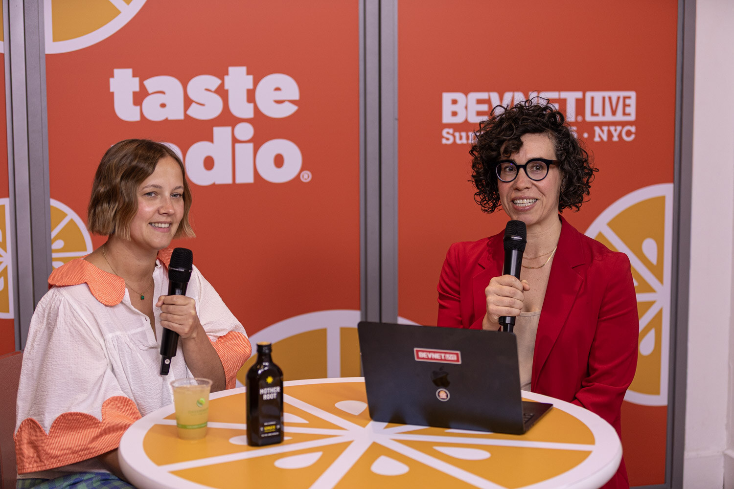 A woman with short hair in a white blouse with orange trim holds a microphone at a table with a Mother Root bottle and a glass.