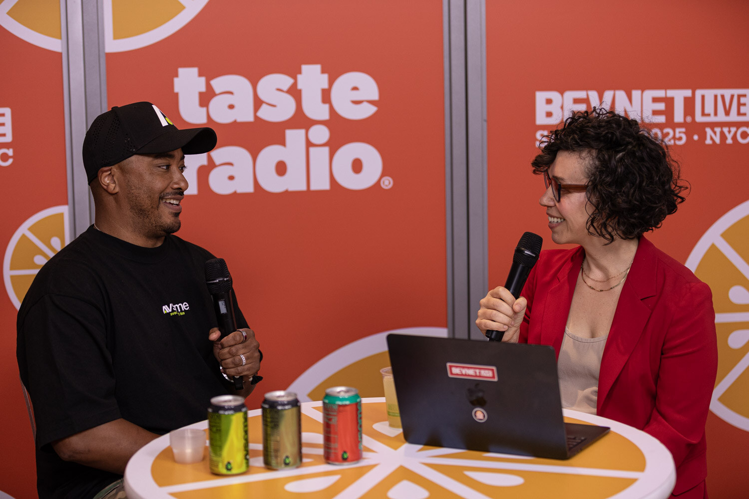 A man in a black cap and AVSOME t-shirt and a woman in a red blazer hold microphones at a table with three cans.