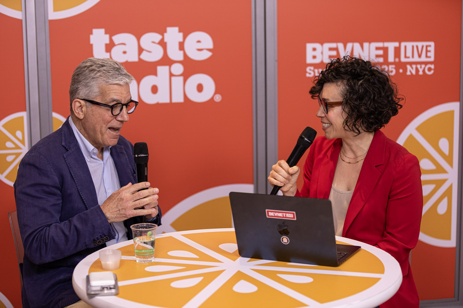 A man with gray hair and glasses in a navy blazer and a woman in a red blazer hold microphones at a table with a laptop.