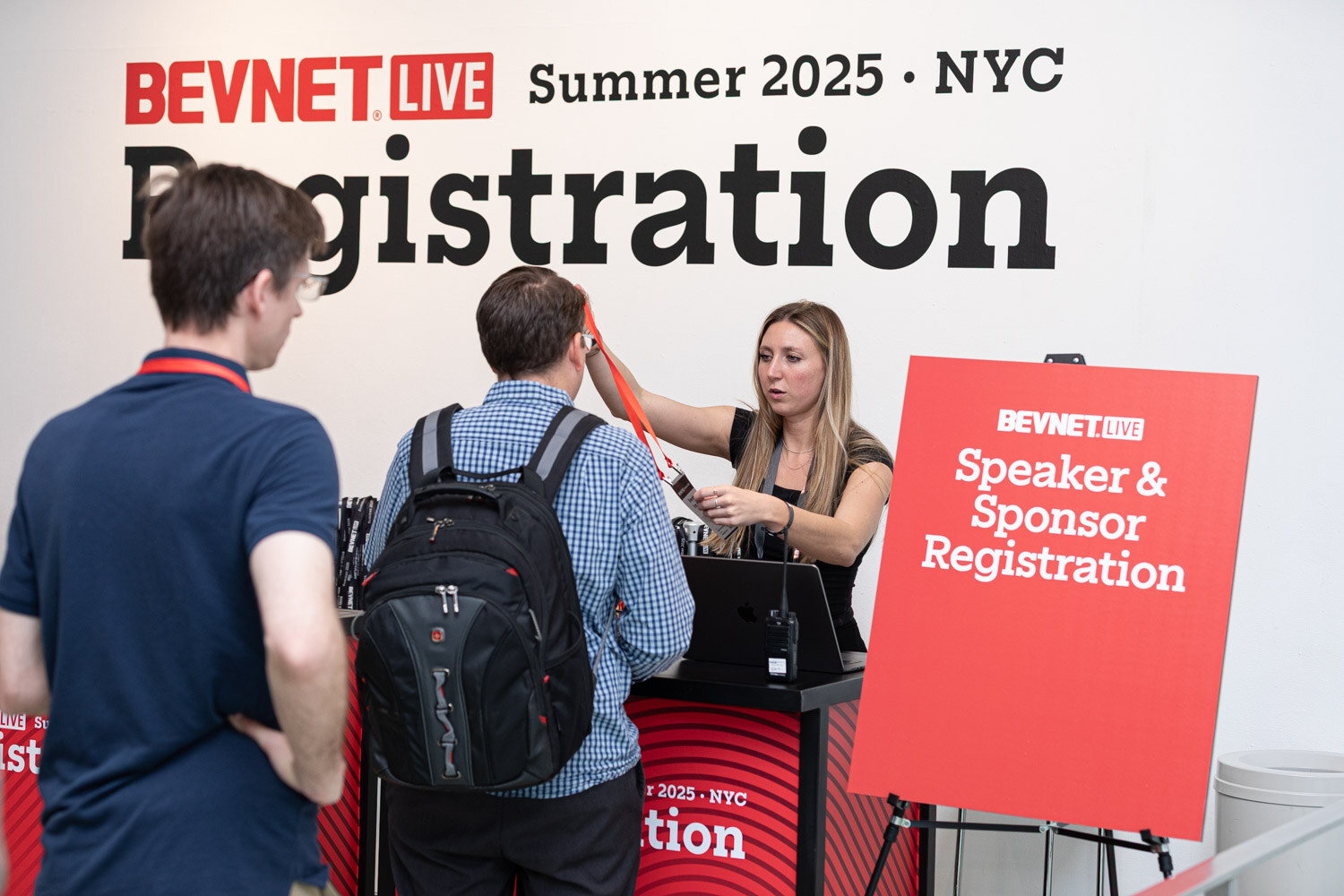Two attendees at a desk with a woman scanning a badge in front of a BevNET Live Registration wall sign.