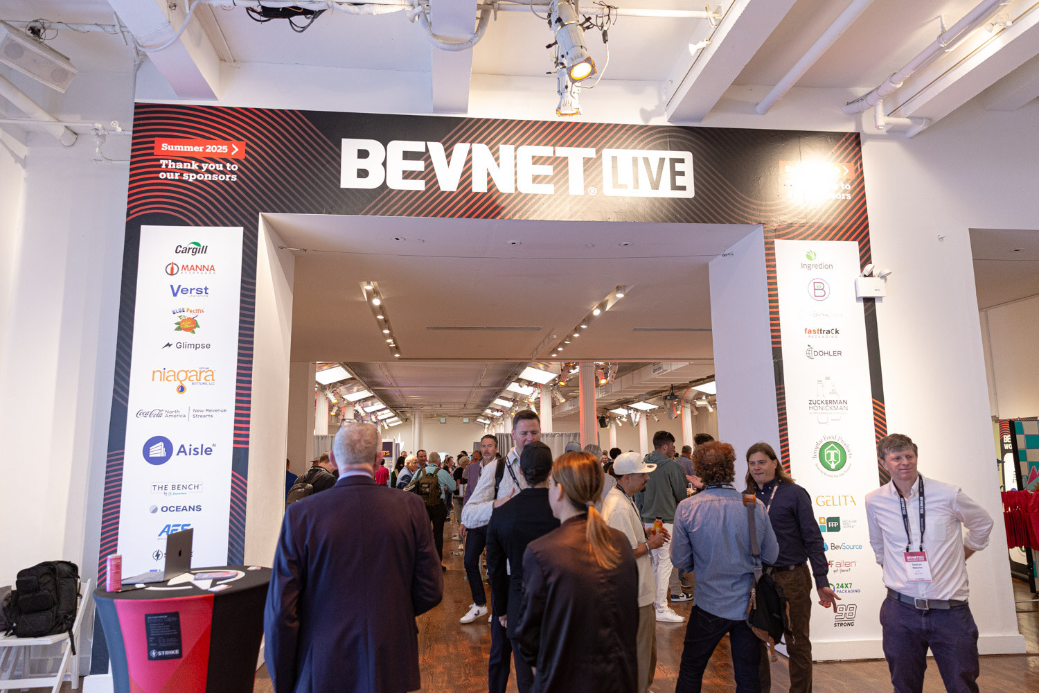A black BevNET Live archway with sponsor logos and attendees walking into a bright exhibit hall.