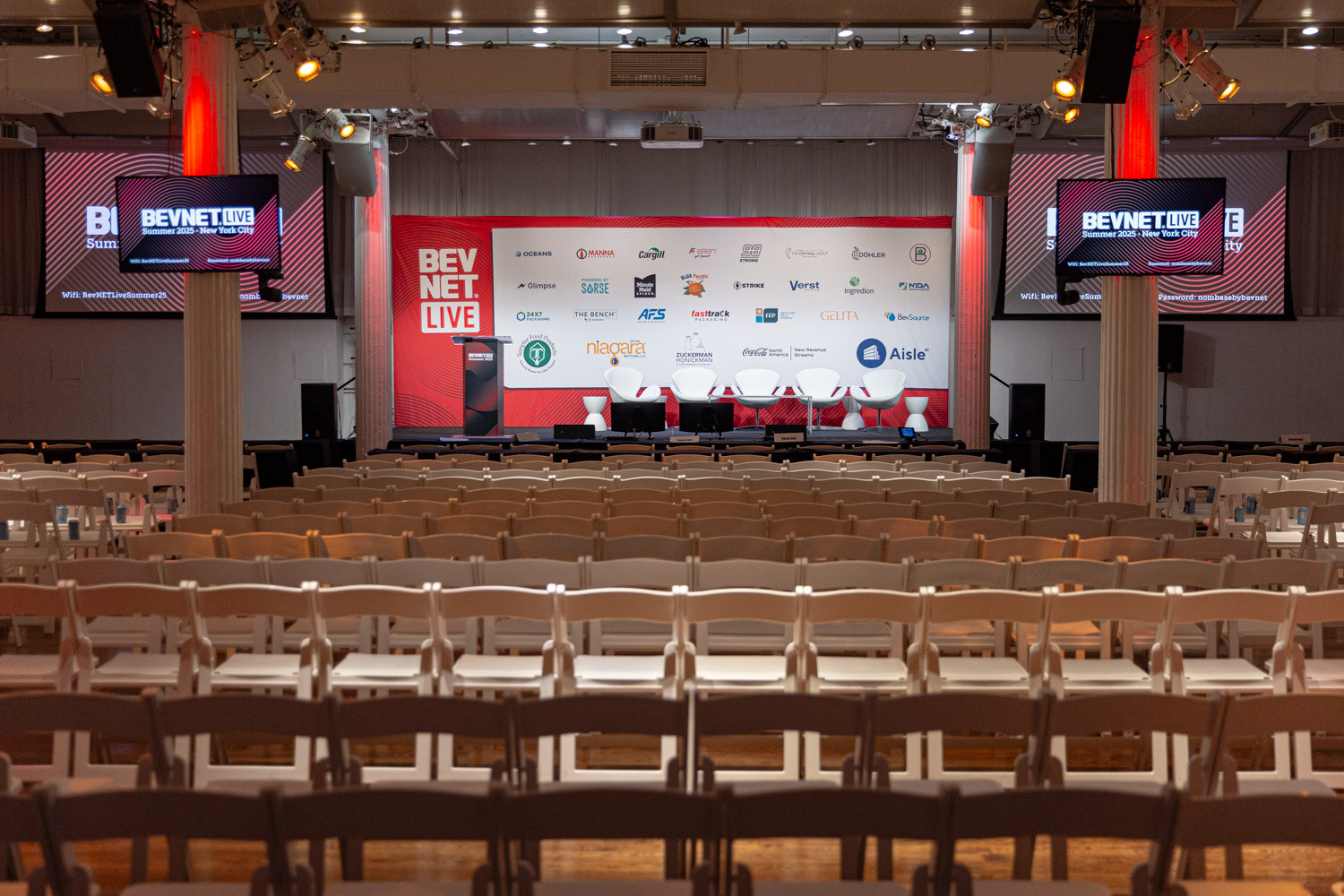 Rows of wooden chairs facing a stage with a red BevNET Live backdrop and screens.