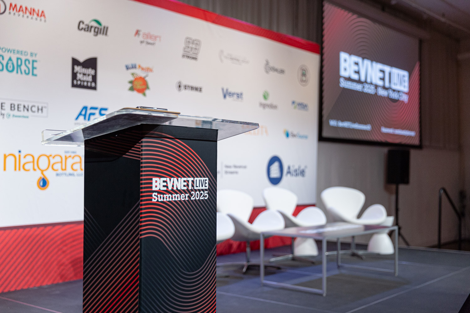 A black podium marked BevNET Live Summer 2025 beside white chairs on a stage in front of a sponsor backdrop.