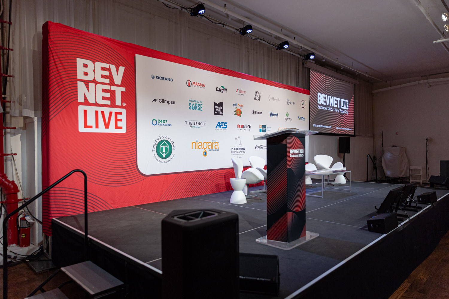 A stage with a red BevNET Live banner, a black podium, white chairs and screens.