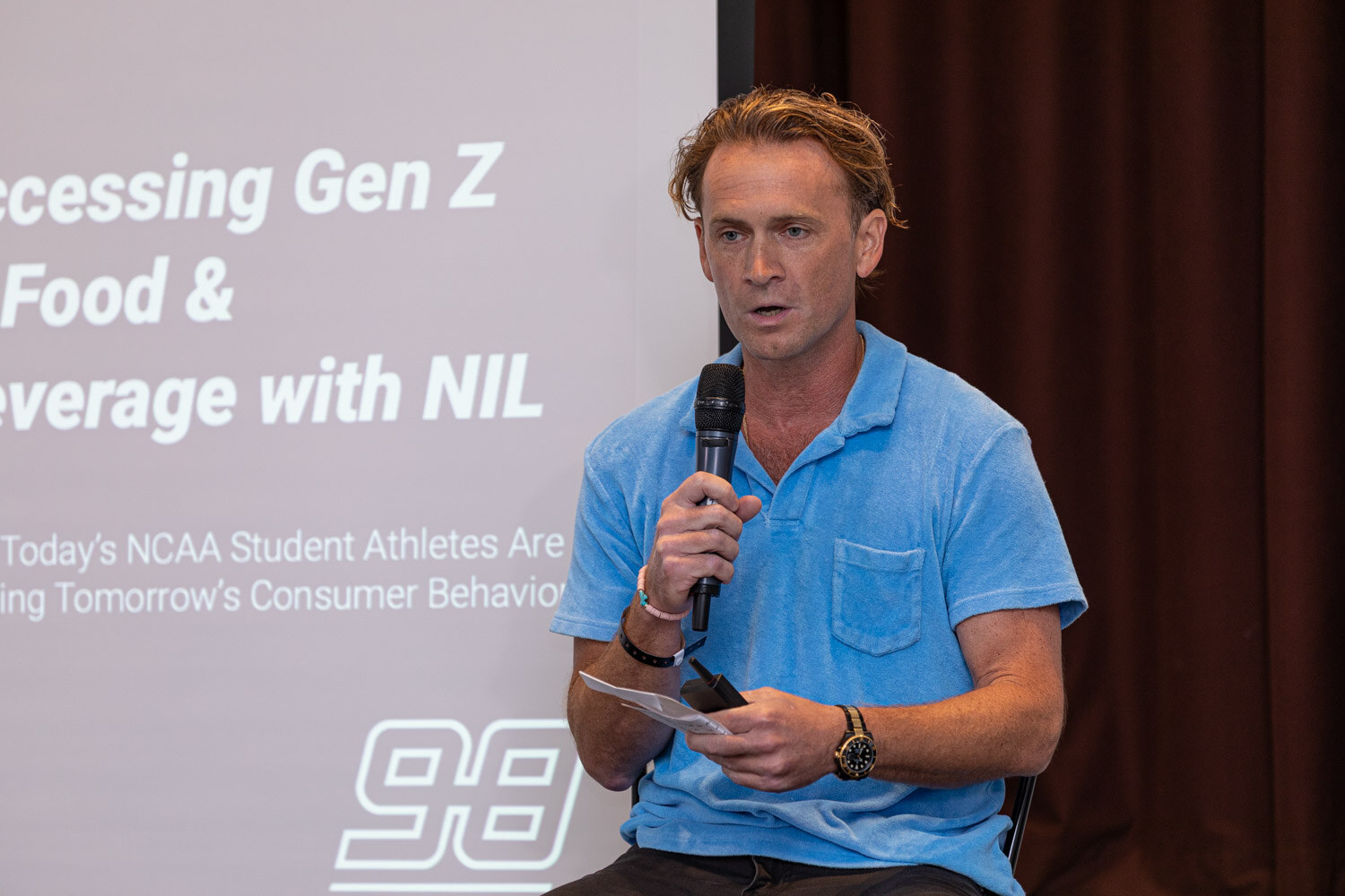 A man in a light blue polo shirt speaks into a microphone in front of a slide reading Accessing Gen Z in Food & Beverage with NIL and a 98 Strong logo.