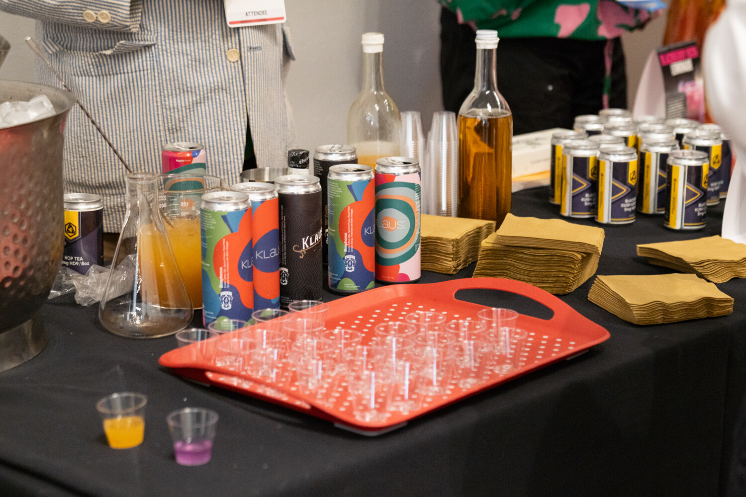A black tablecloth holding canned beverages, bottles of juice and a red tray of small sample cups.