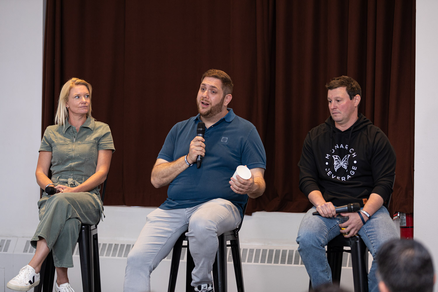 Two men and a woman sit on stools in front of a dark curtain, each with a microphone.