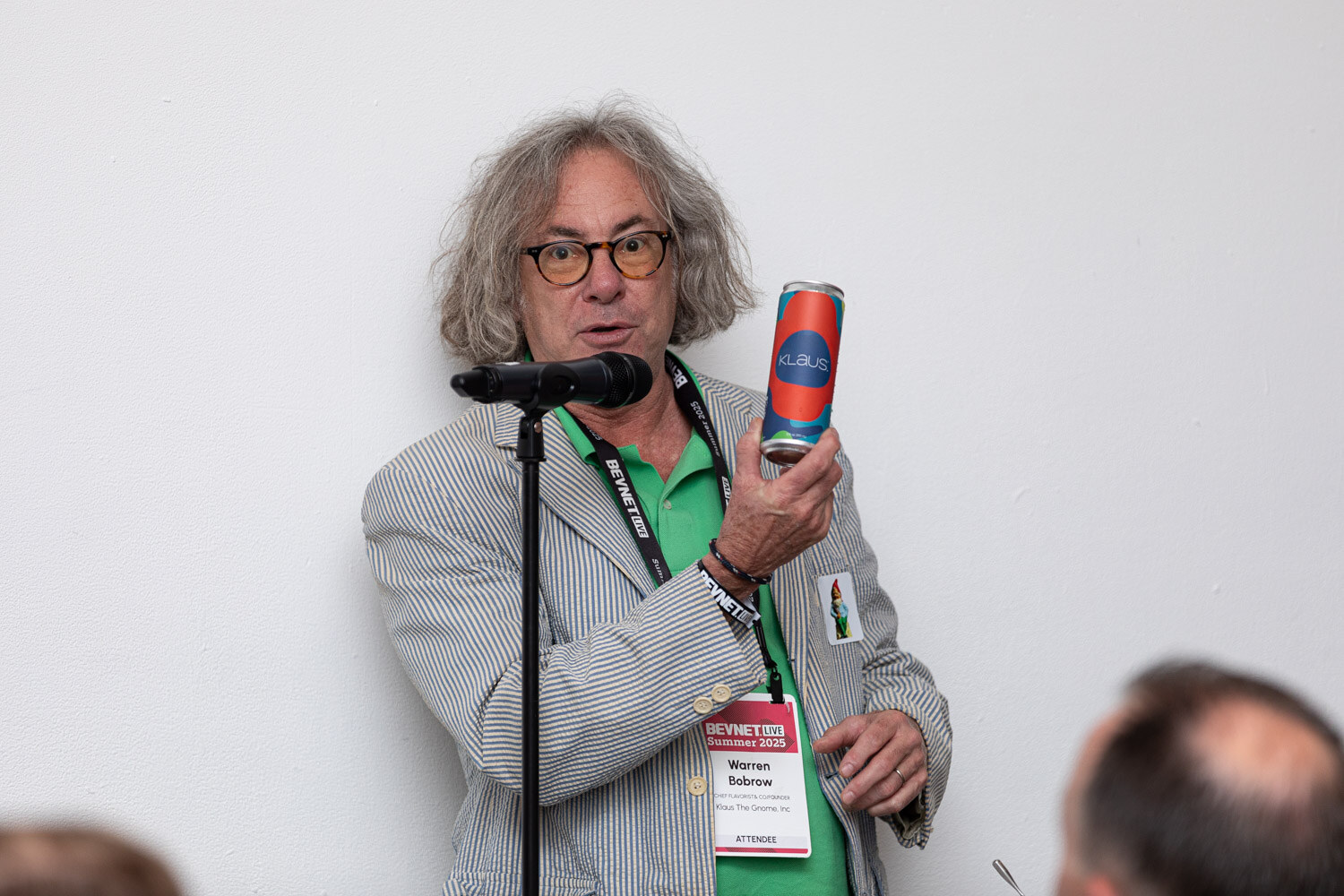 A man with long gray hair and glasses in a striped jacket holds a canned beverage up at a standing microphone.