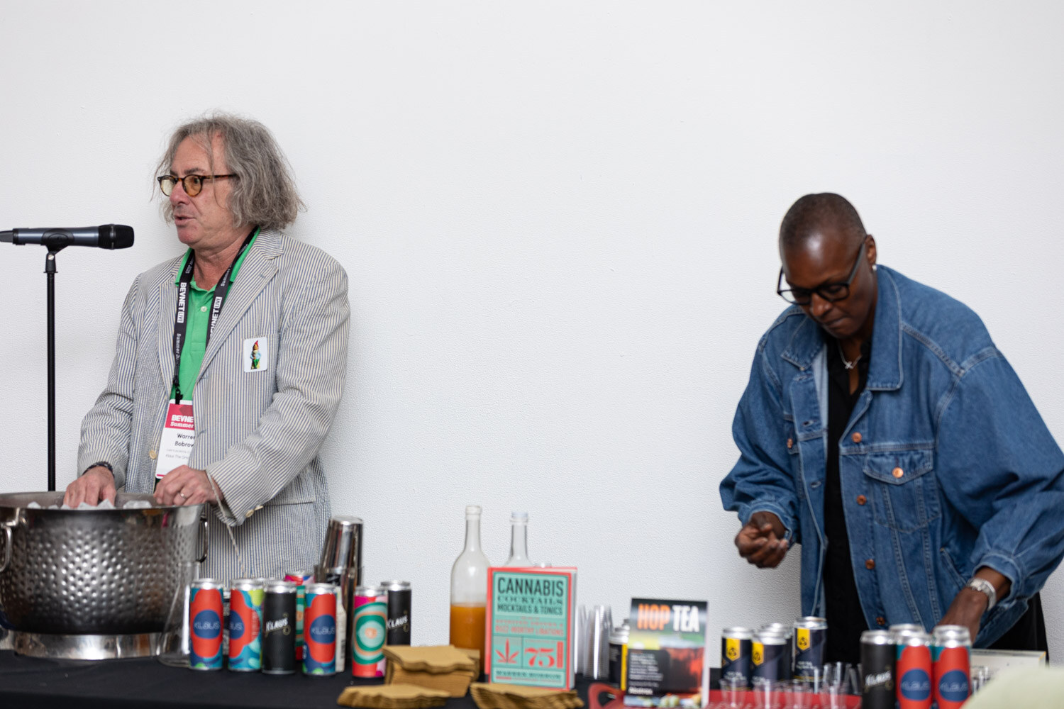 Two men stand behind a table with a metal ice bucket, canned beverages and a Cannabis Cocktails and Tonics book.