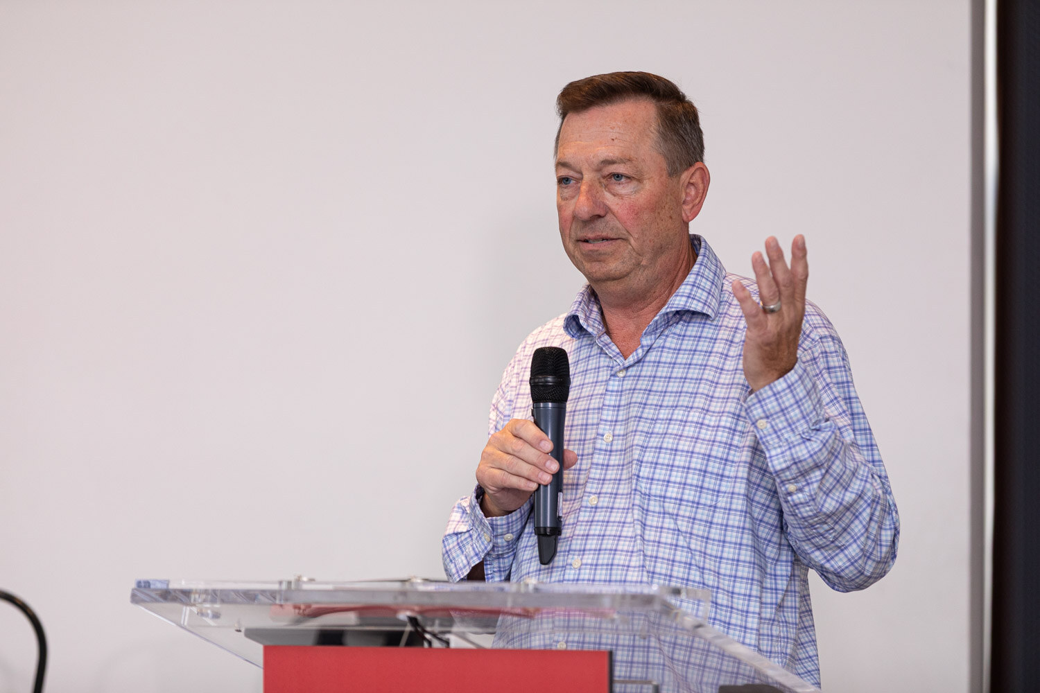 A man in a blue checked shirt holds a microphone in front of a white screen and dark curtain.