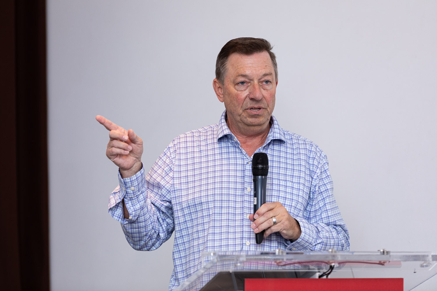 A man in a blue checked shirt points while speaking into a microphone at a podium.