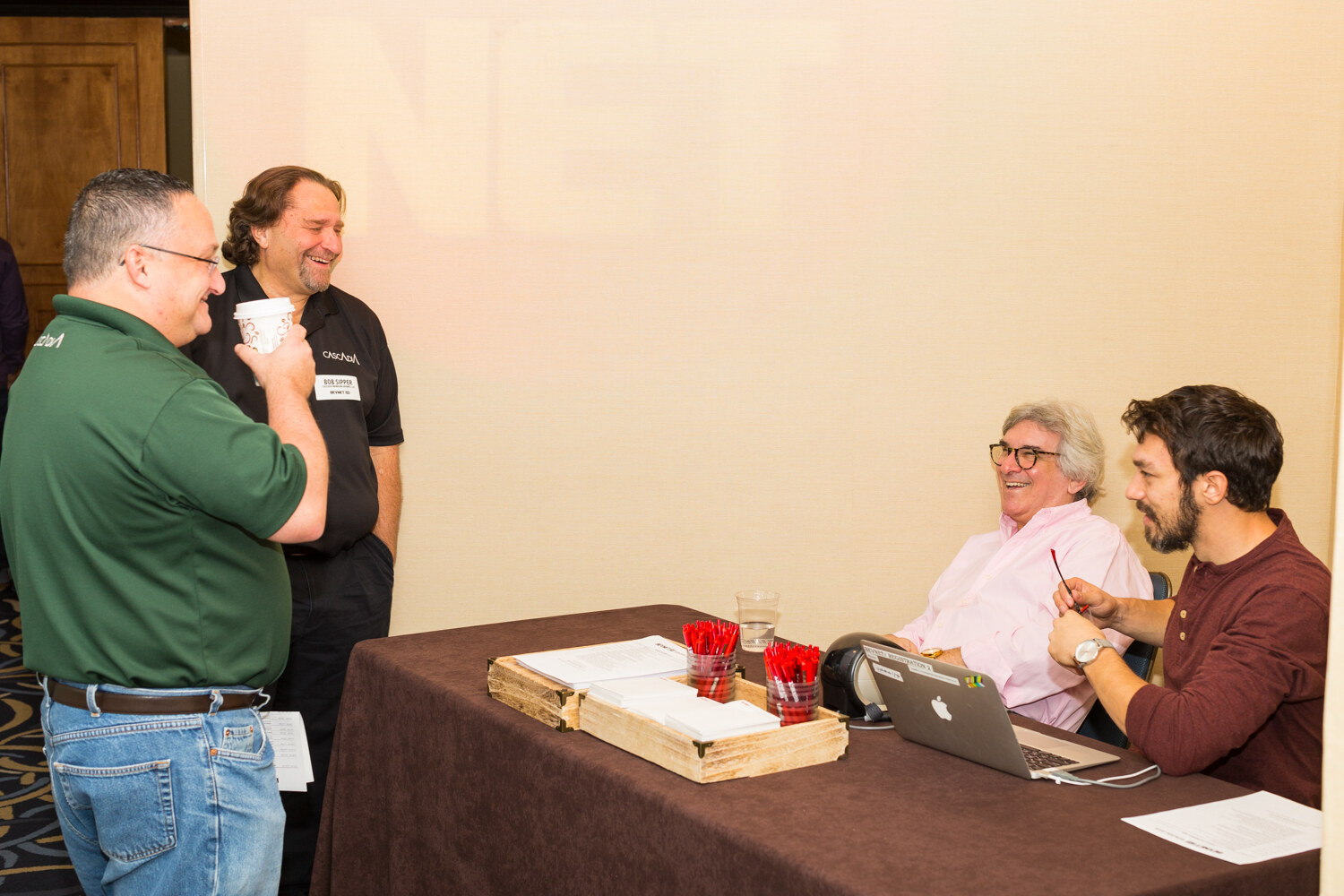 Two men stand and chat, one in a green polo holding a coffee cup, while two seated men behind a brown-draped table with red pens, name-badge stock and a laptop laugh.