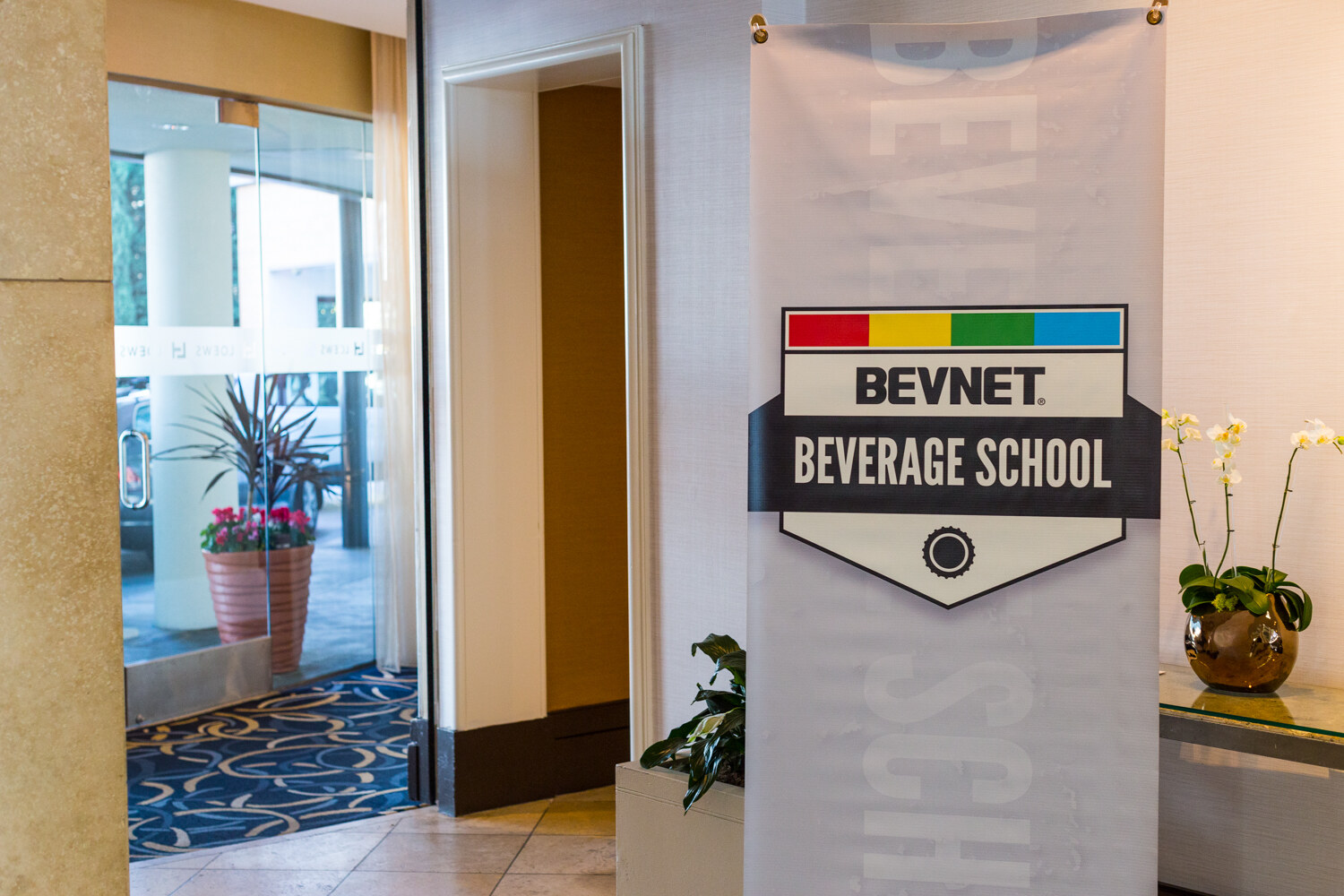 Tall gray banner with the BevNET Beverage School badge logo standing beside a doorway, with a glass door at left and a vase of white orchids at right.