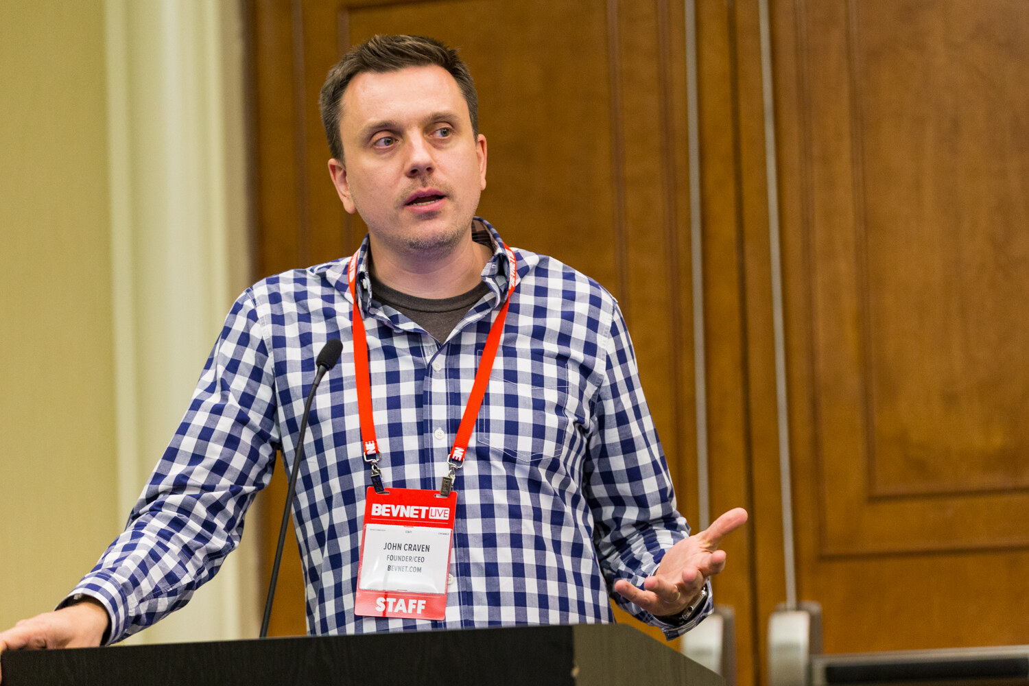 A man in a blue and white gingham shirt with a red BevNET Live staff lanyard speaks at a black lectern in front of wooden doors, one hand open.