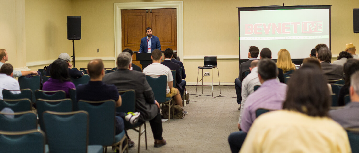 View from the back of a hotel meeting room: rows of seated attendees face a man at a lectern and a projection screen showing the BevNET Live logo.