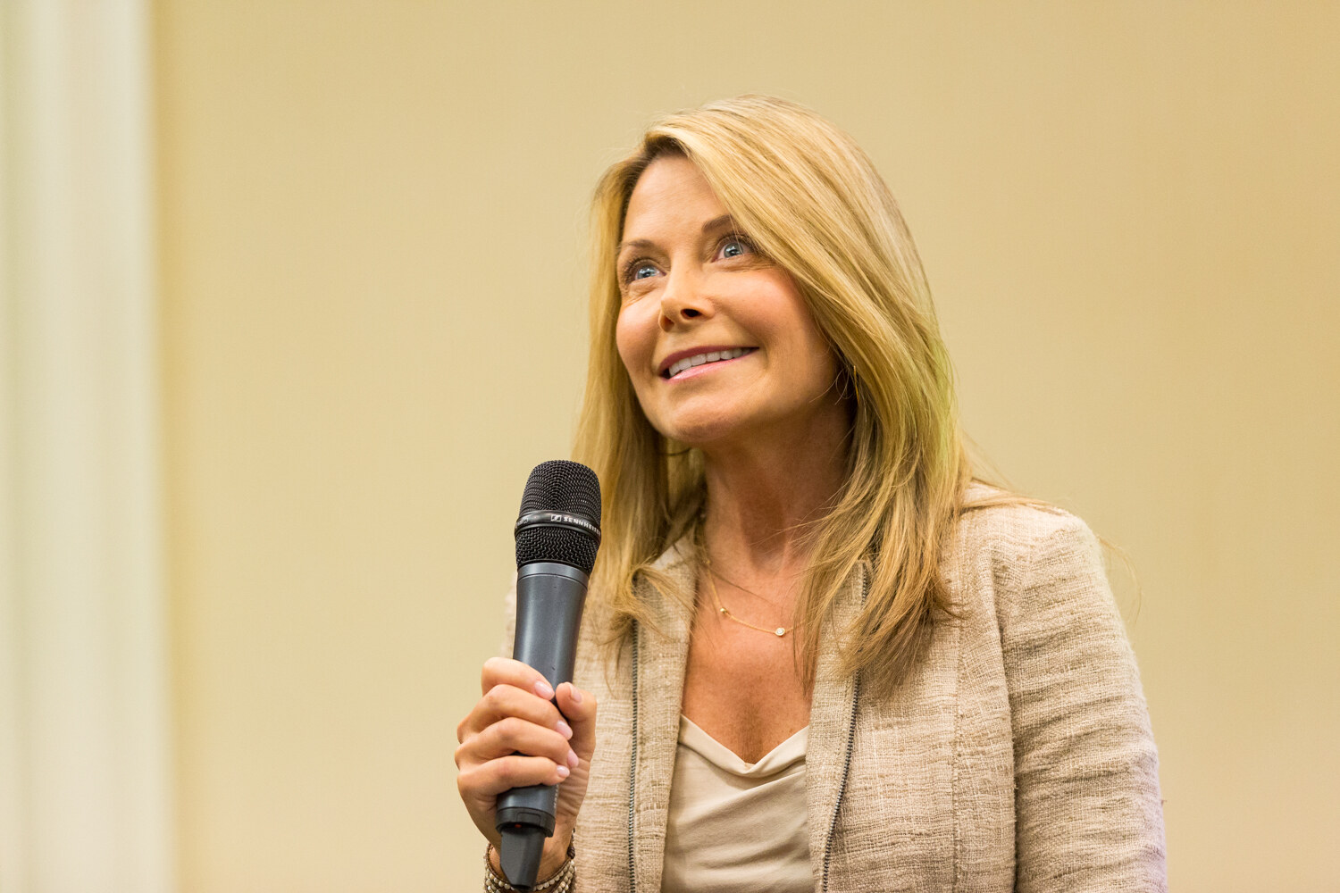 Close view of a smiling woman with long blond hair in a beige jacket holding a black microphone and looking up and to the side.