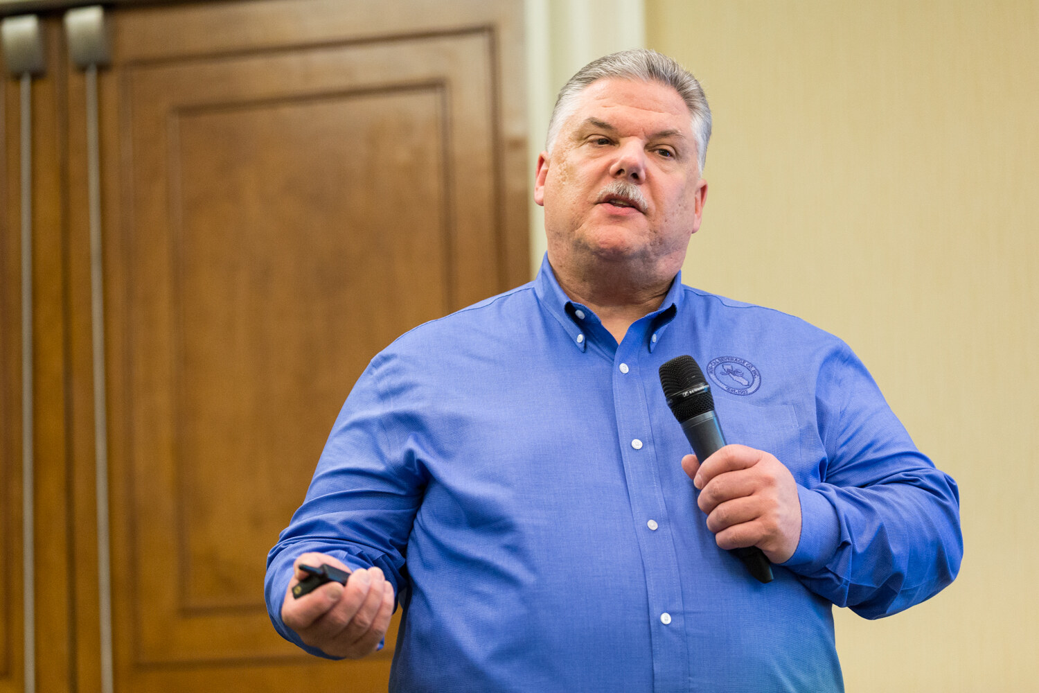 A gray-haired man with a mustache in a blue shirt holds a handheld microphone in one hand and a slide clicker in the other in front of wooden doors.