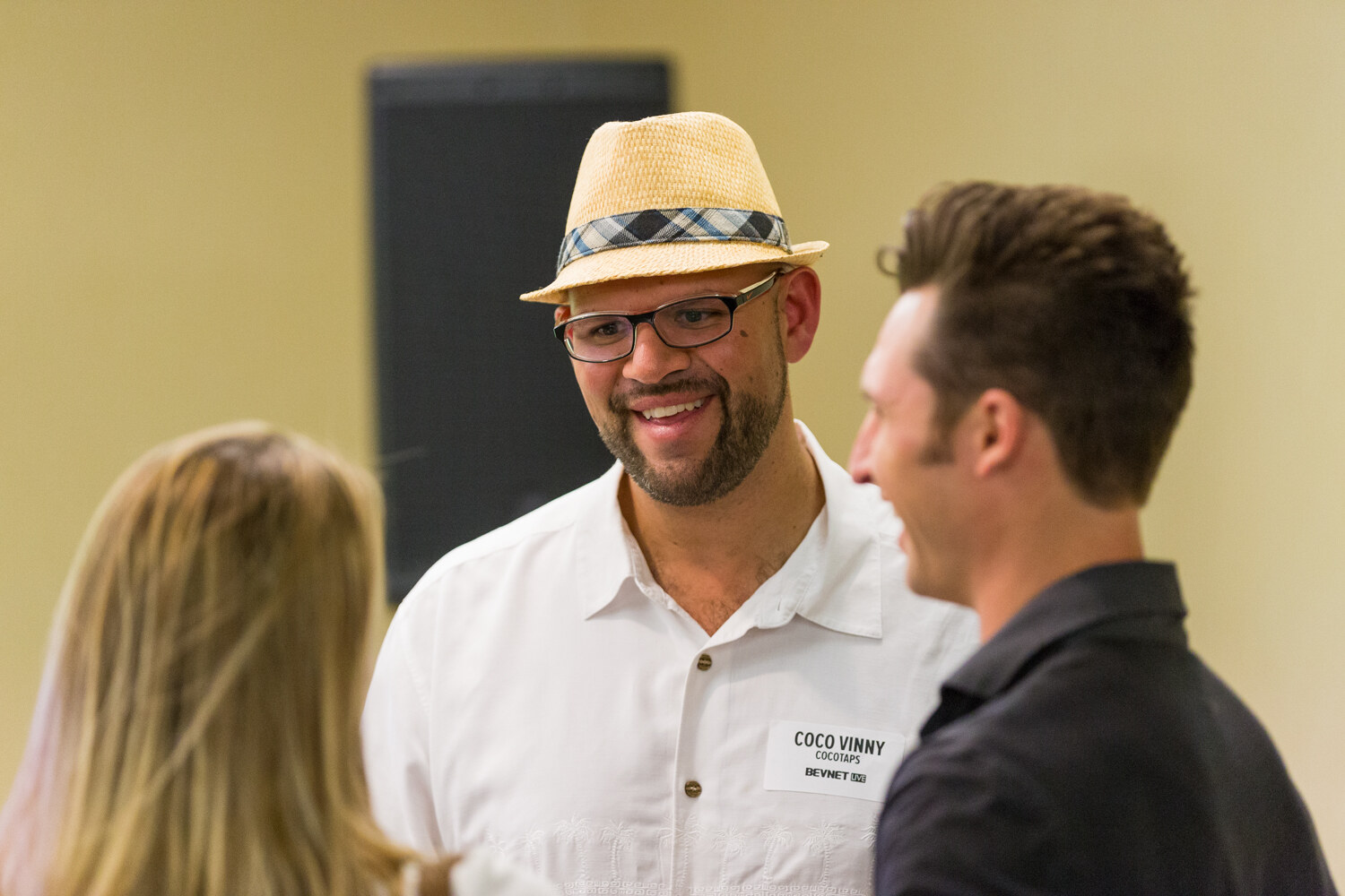 A smiling bearded man in a straw fedora, glasses and a white shirt with a Cocotaps name badge talks with a woman seen from behind and a man in a dark shirt in profile.