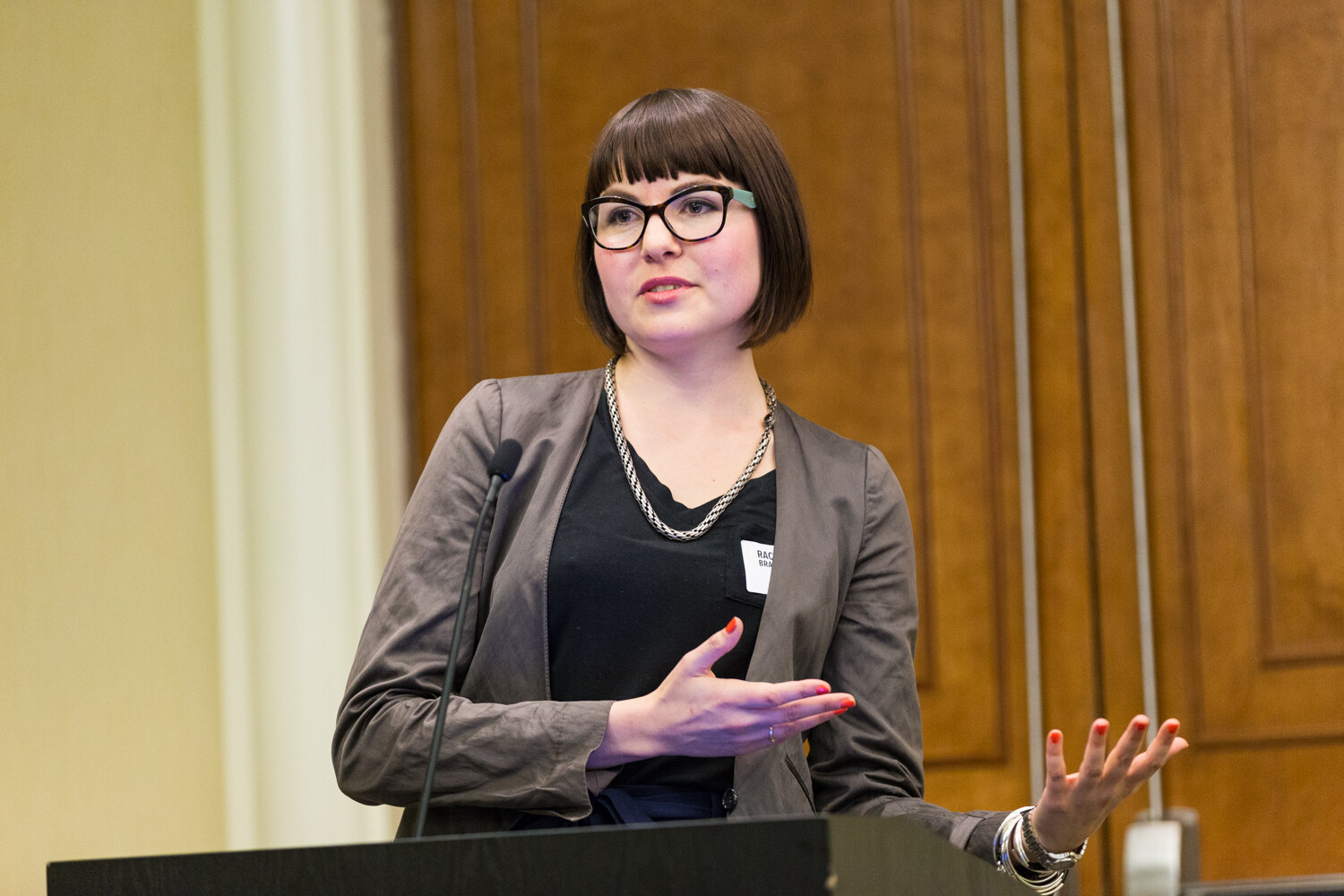 A woman with a dark bob haircut and black-framed glasses, wearing a gray jacket and a chain necklace, gestures with both hands at a black lectern.