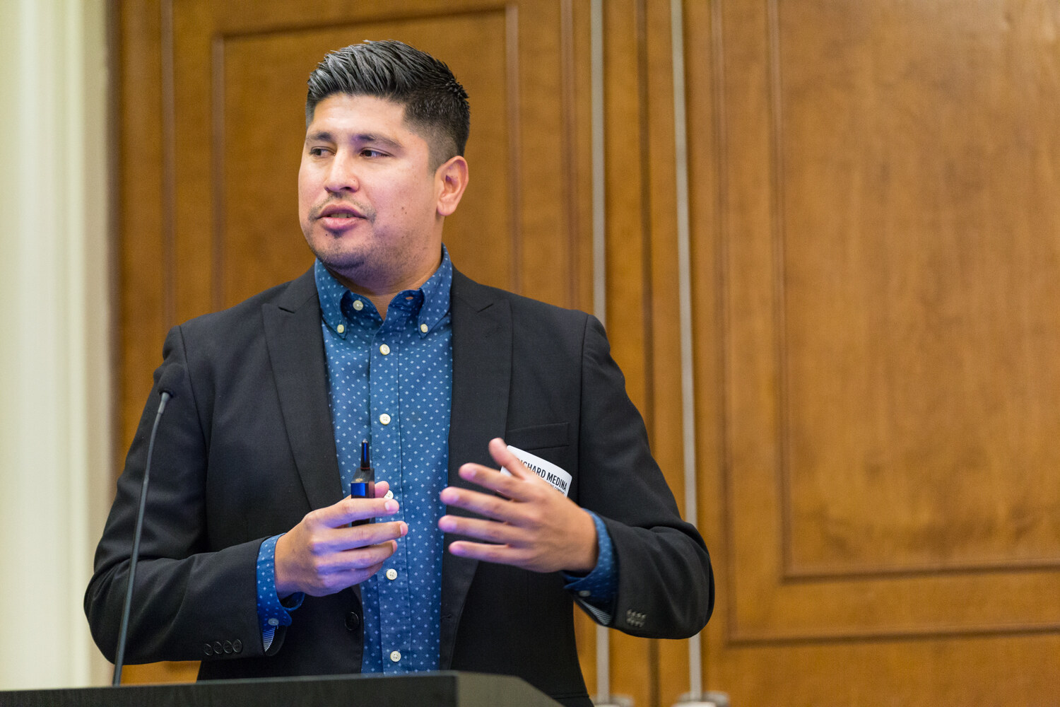 A man in a black blazer and blue polka-dot shirt with a name tag holds a slide clicker and gestures near a lectern microphone in front of wood-paneled doors.