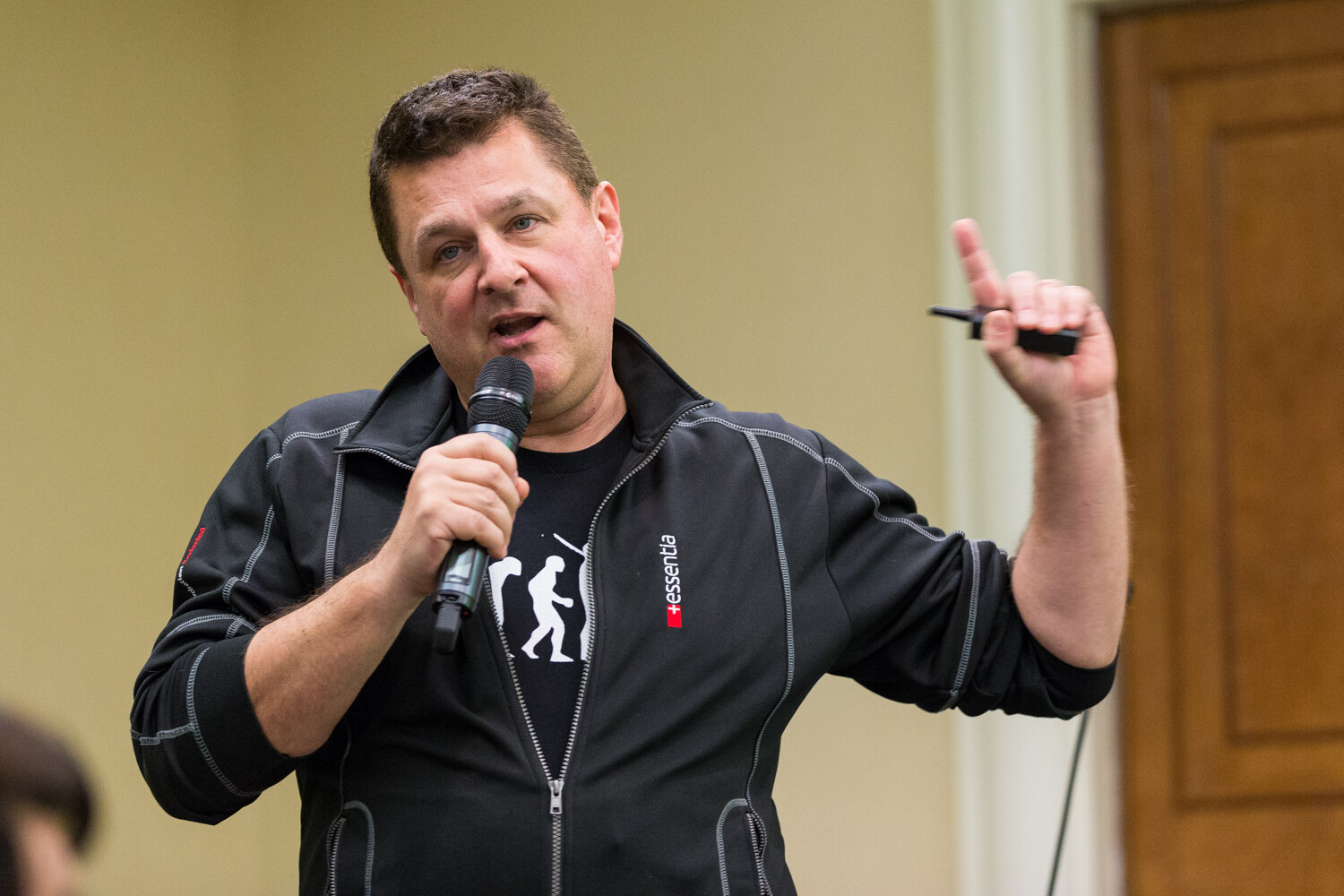 A man in a black zip jacket with an essentia logo holds a microphone in one hand and raises a slide clicker in the other.