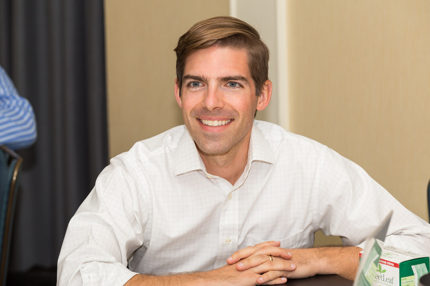 A smiling man in a white checked shirt sits with his hands clasped at a table; a green SweetLeaf box is partly visible at the lower right.
