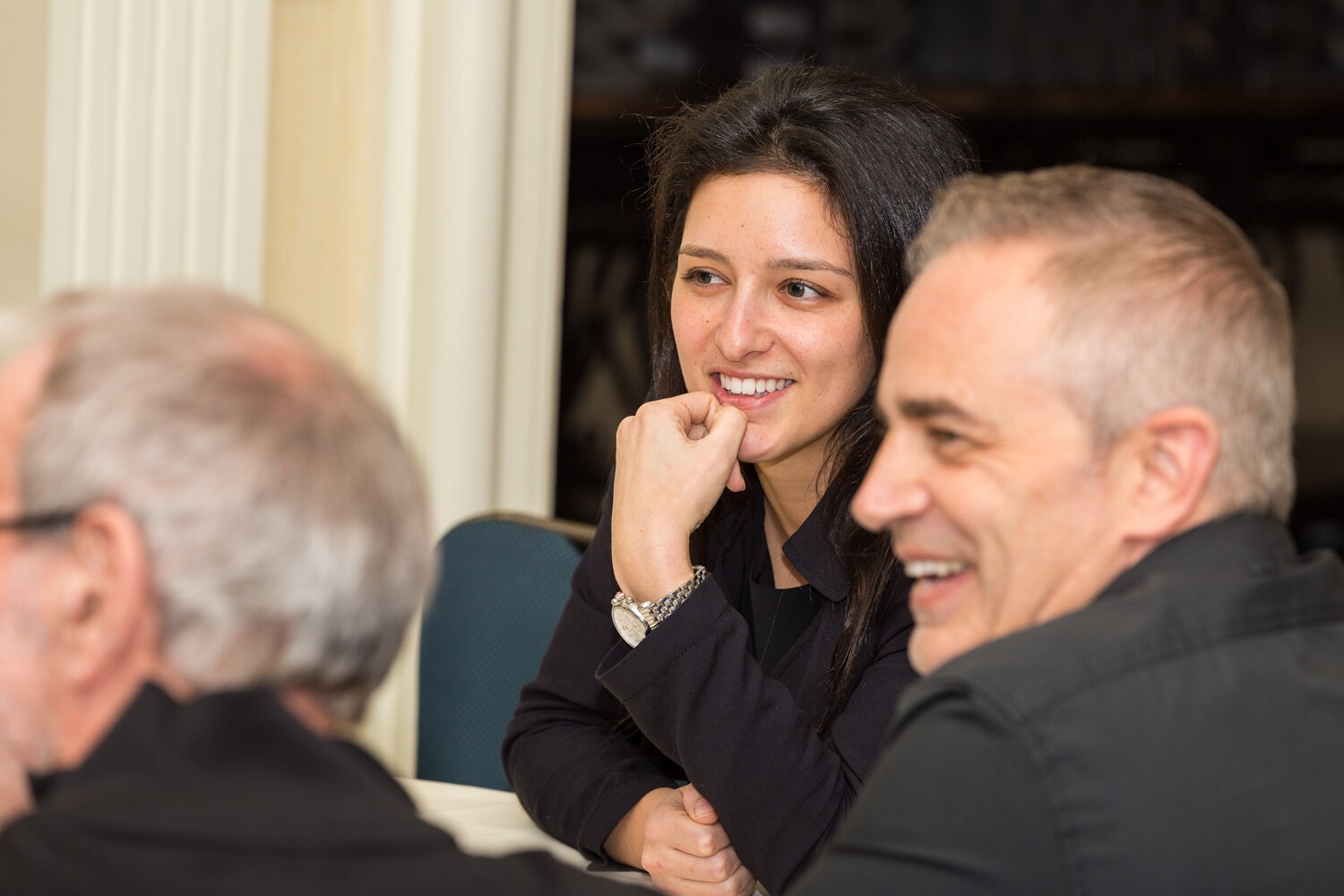 A dark-haired woman in a black shirt rests her chin on her fist and smiles beside a laughing gray-haired man; another man is blurred in the foreground.