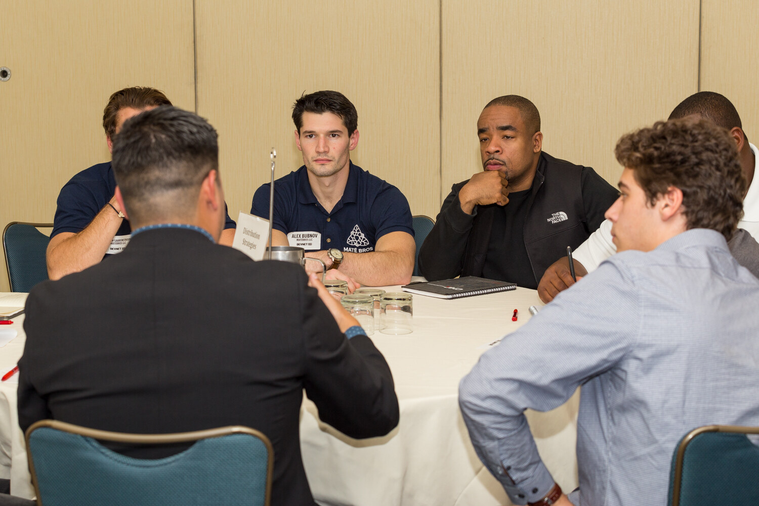 Five people sit around a white-clothed round table with a card reading Distribution Strategies; one man wears a navy Mate Bros polo and two others face away from the camera.