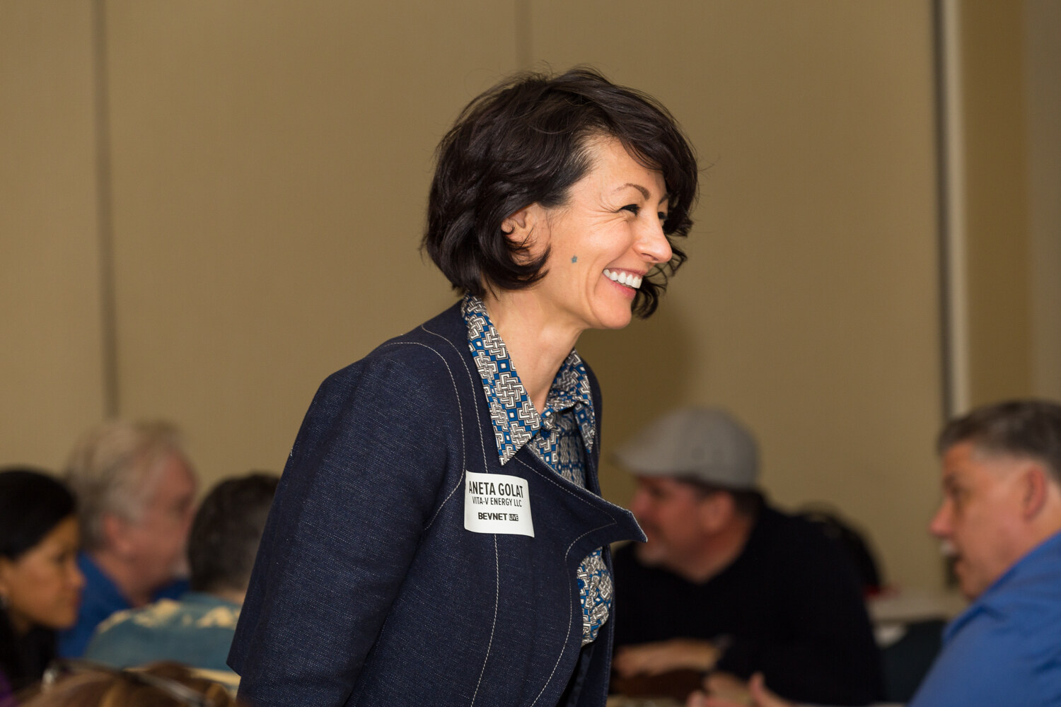Side view of a laughing woman with short dark hair in a navy denim-style jacket and a patterned collar, with seated attendees blurred behind her.