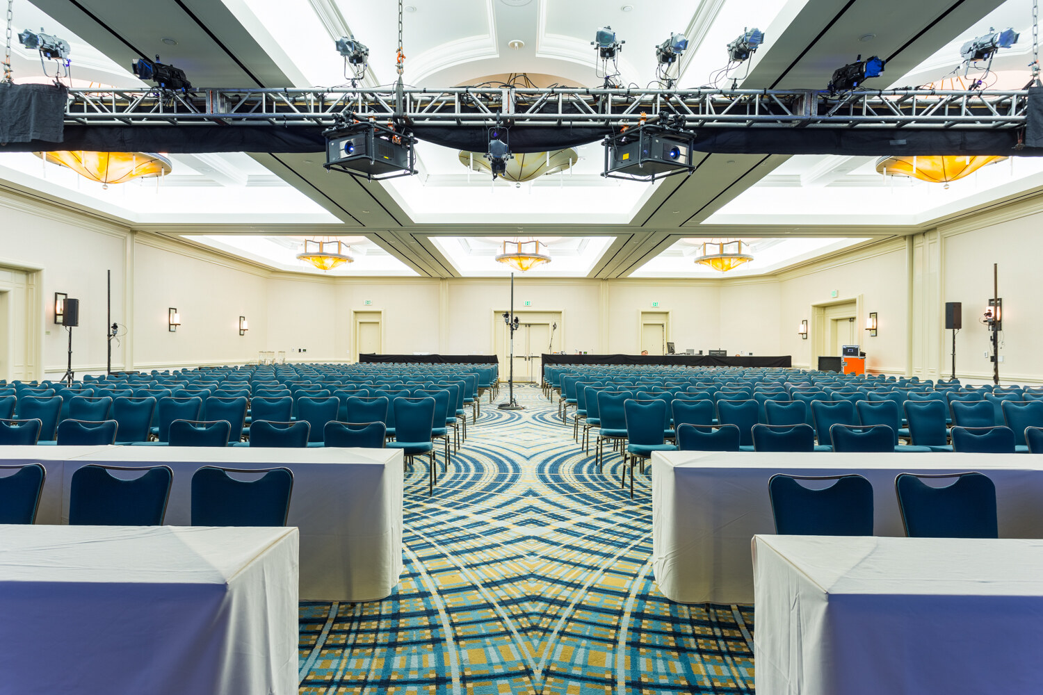 Wide view down a carpeted center aisle of a hotel ballroom with rows of teal chairs, white-draped tables in the foreground, ceiling truss with lights and projectors and a far stage.