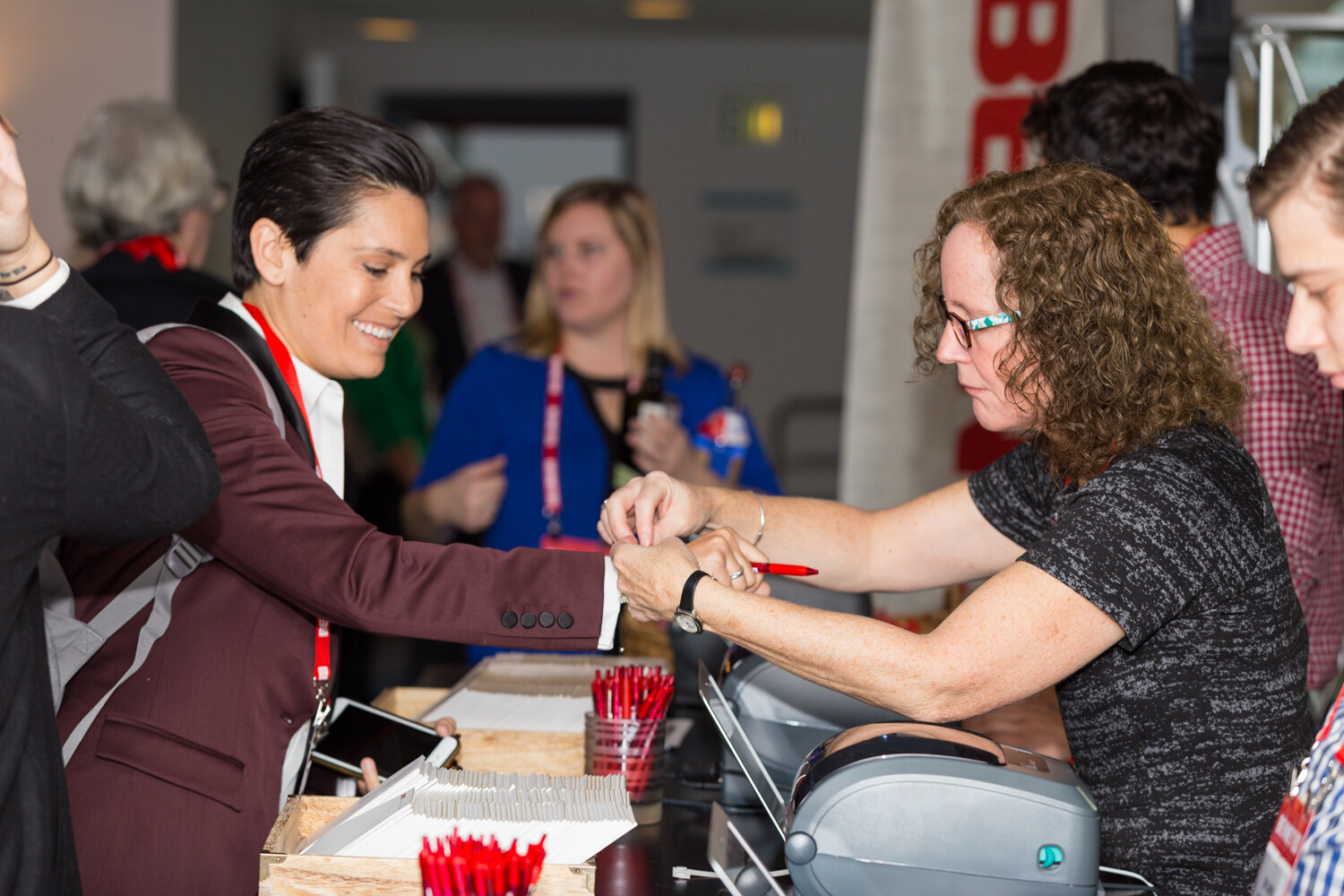 A smiling woman in a maroon blazer holds out her wrist while a woman with curly hair and glasses fastens a wristband, beside a desk with red pens, name badge stock and a badge printer.