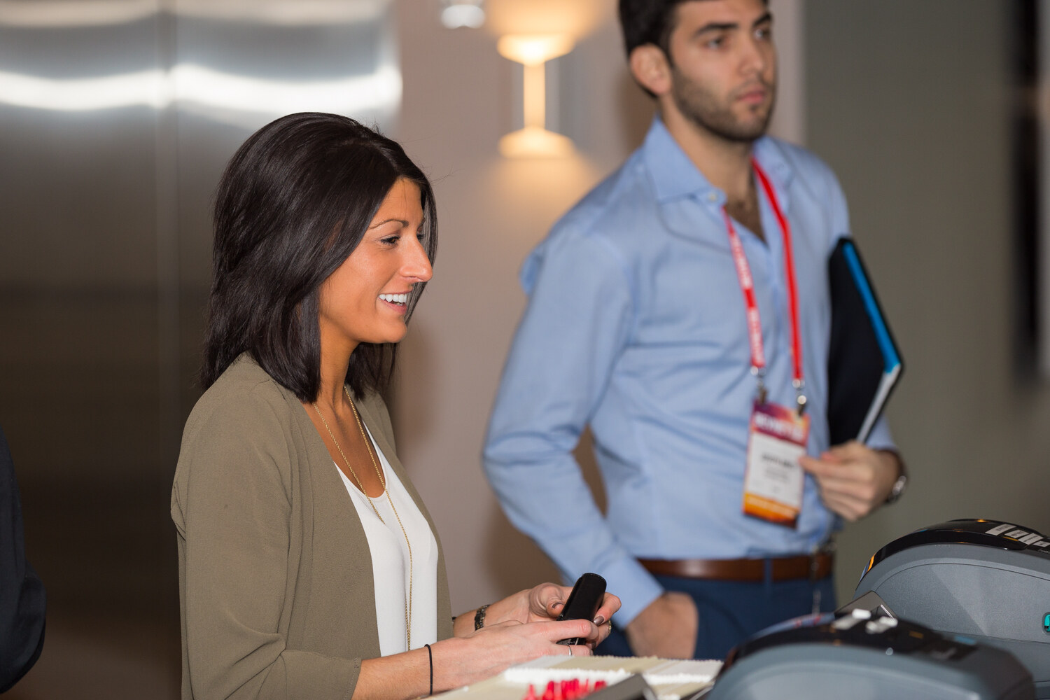 A woman with dark hair and an olive cardigan smiles over a desk with a badge printer; behind her a man in a light blue shirt with a BevNET Live lanyard holds a folder.