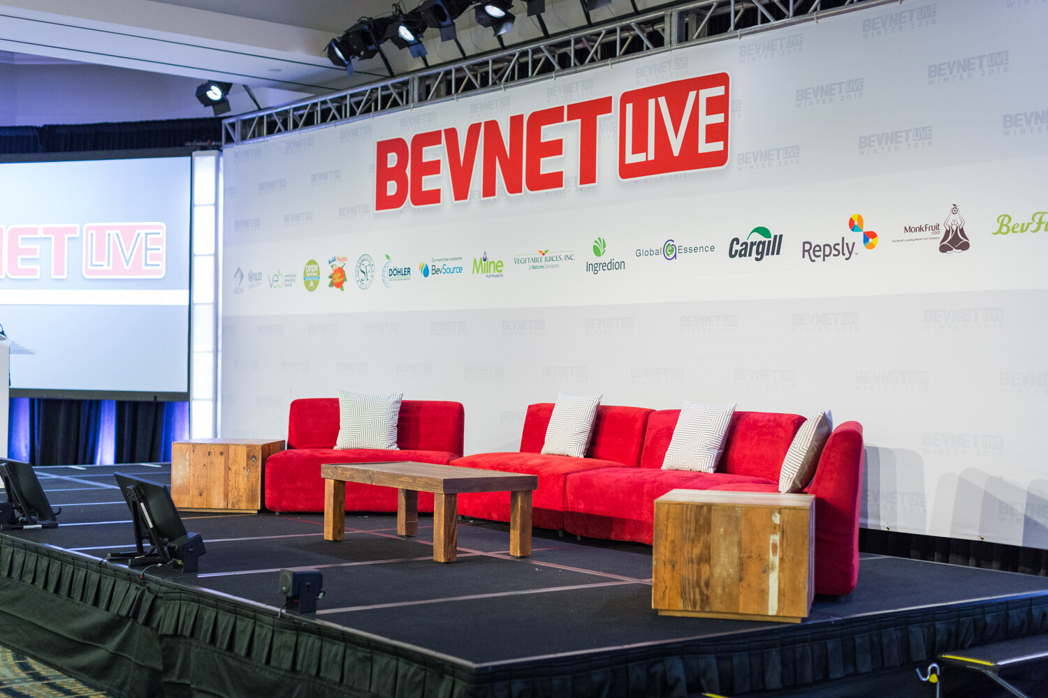 A raised black stage with two red couches, a wooden coffee table and side tables in front of a white backdrop with a large red BEVNET LIVE logo and sponsor logos including Cargill, Repsly and Monk Fruit.