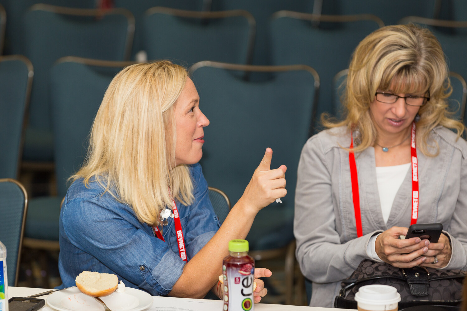 A blond woman in a denim shirt points with her thumb while a woman in a gray jacket looks at her phone; a bottle of cherry juice, a bread roll and a coffee cup sit on the table.