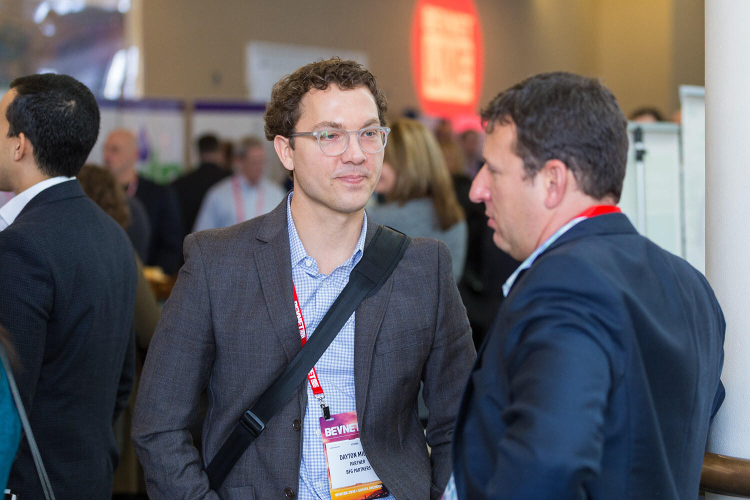 A man with curly hair, clear glasses, a gray blazer and a crossbody bag looks on while a man in a navy suit talks; other attendees are blurred behind.