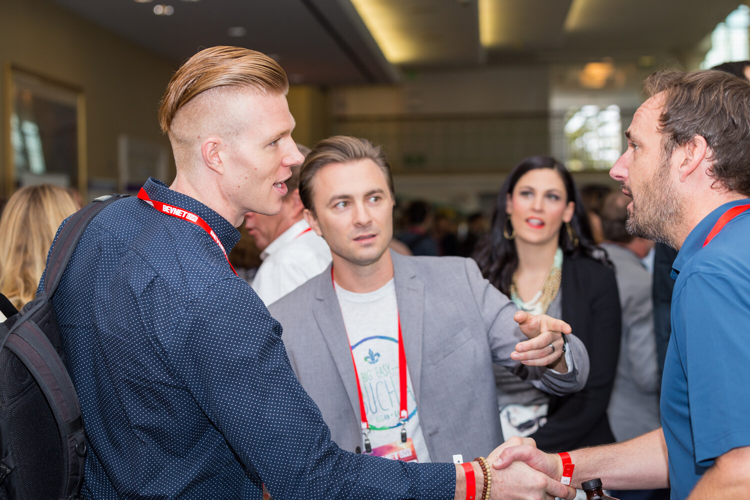 A tall man with a blond undercut and a backpack shakes hands with a bearded man in a blue polo while a man in a gray blazer and a woman in black watch.