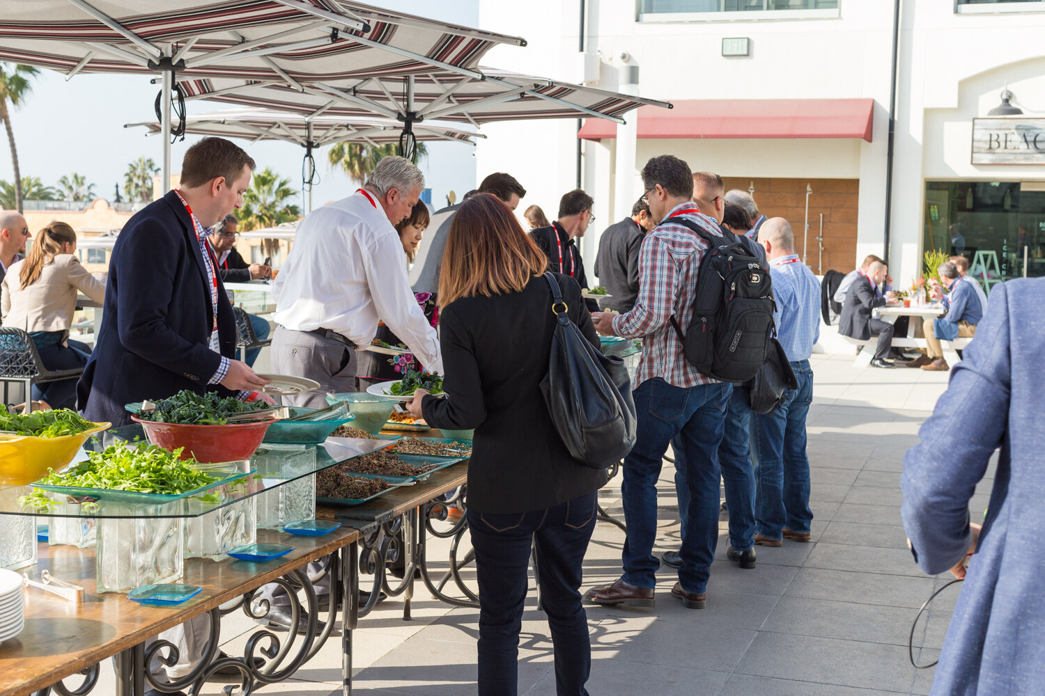 Attendees with red lanyards line up along a buffet of salads and greens on a sunny terrace under striped umbrellas, with palm trees in the background.