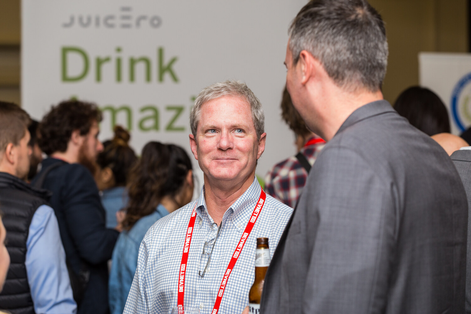A gray-haired man in a checked shirt with a red BevNET lanyard smiles up at a man in a gray blazer seen from behind, in front of a Juicero Drink Amazing Juice banner.
