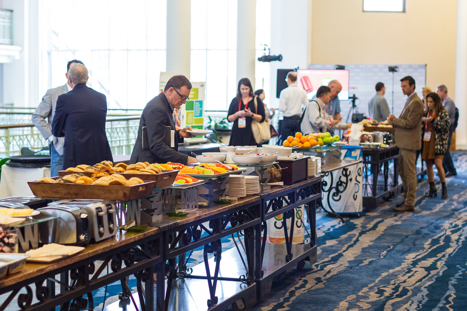 A buffet table with a tray of pastries, toasters and fruit while attendees in business casual clothes queue and serve themselves in a bright atrium with a blue patterned carpet.