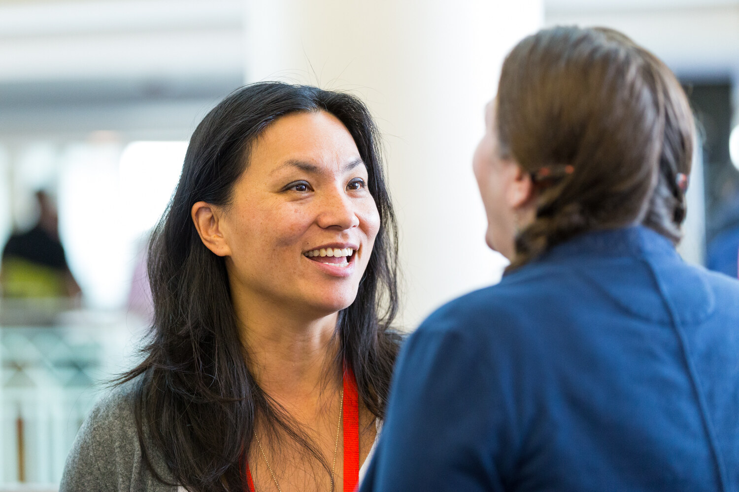 A smiling woman with long dark hair and a red lanyard faces a person in a navy jacket seen from behind, with a bright atrium blurred behind them.