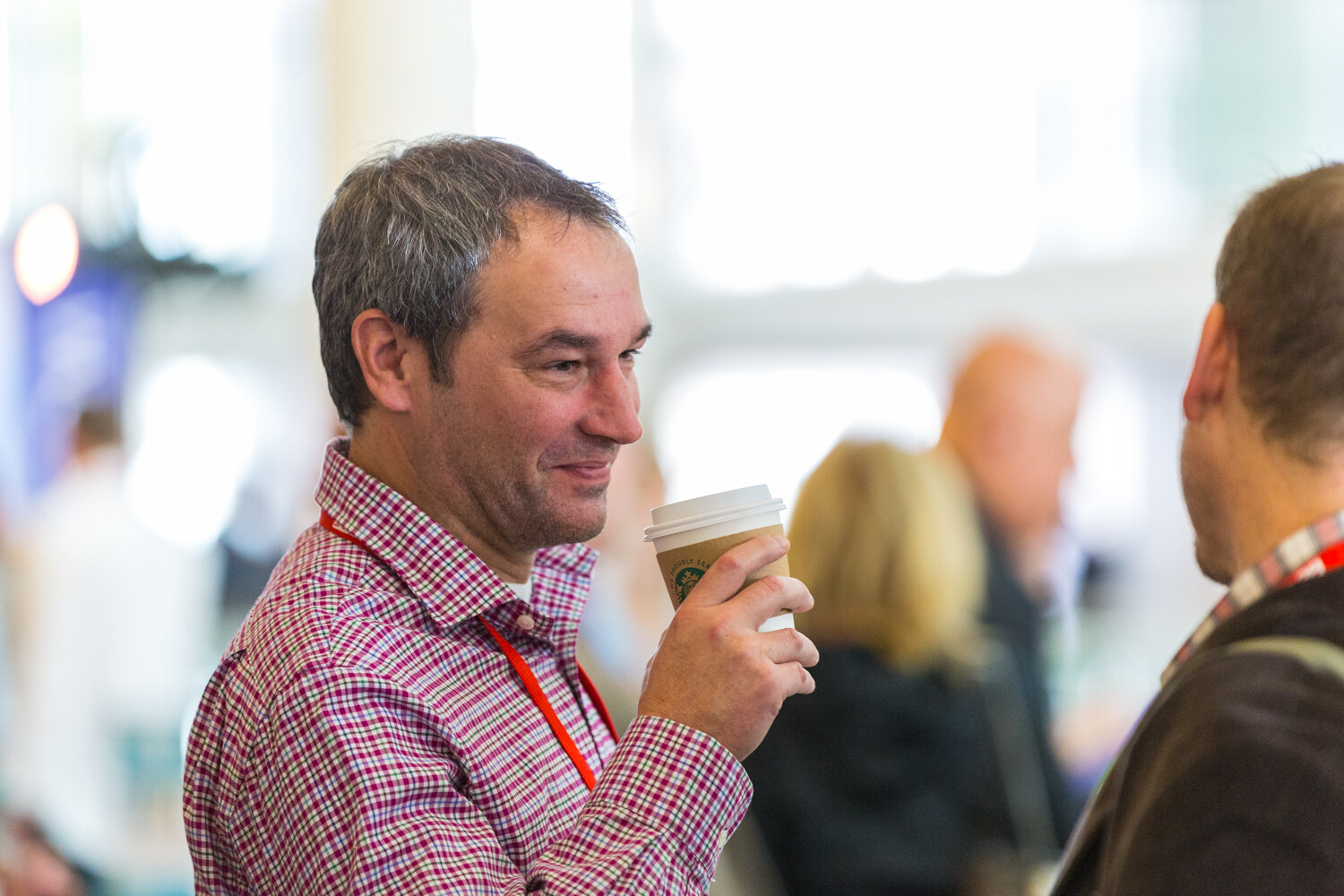 A gray-haired man in a red and white checked shirt with a red lanyard holds a paper coffee cup and smiles as he talks with another attendee seen from behind.
