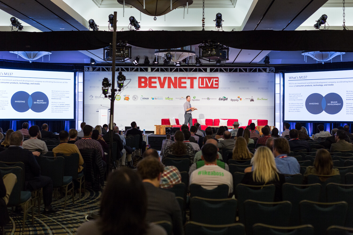 View from the back of a darkened hotel ballroom: rows of attendees face a man presenting on stage in front of a BEVNET LIVE backdrop, with a slide titled What's M13 on the side screens.