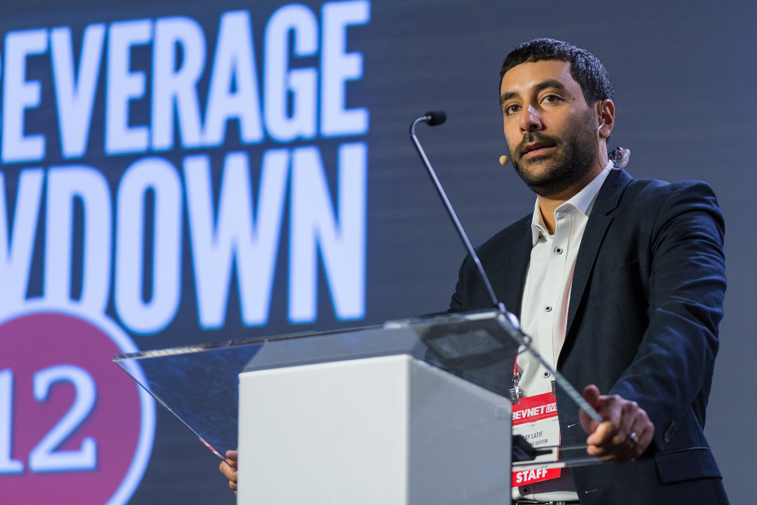 A man in a dark blazer with a headset microphone and a staff badge stands behind a clear lectern with a large screen reading Beverage Showdown 12 behind him.