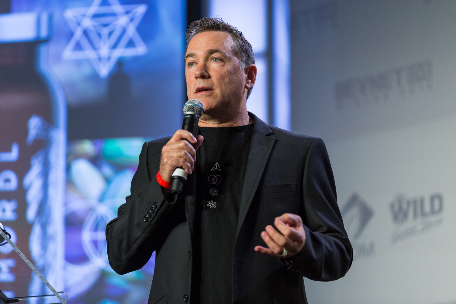 A man in a black blazer and black T-shirt with small symbols holds a microphone while a large screen behind him shows a tall bottle with winged artwork and geometric symbols.