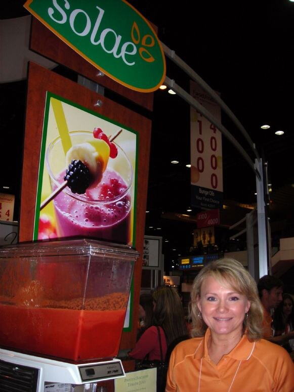 Annette Higgins of Solae, smiling in an orange polo shirt, stands beside a Solae sign, a smoothie photo and a dispenser of red drink, with a Bunge Oils banner behind.