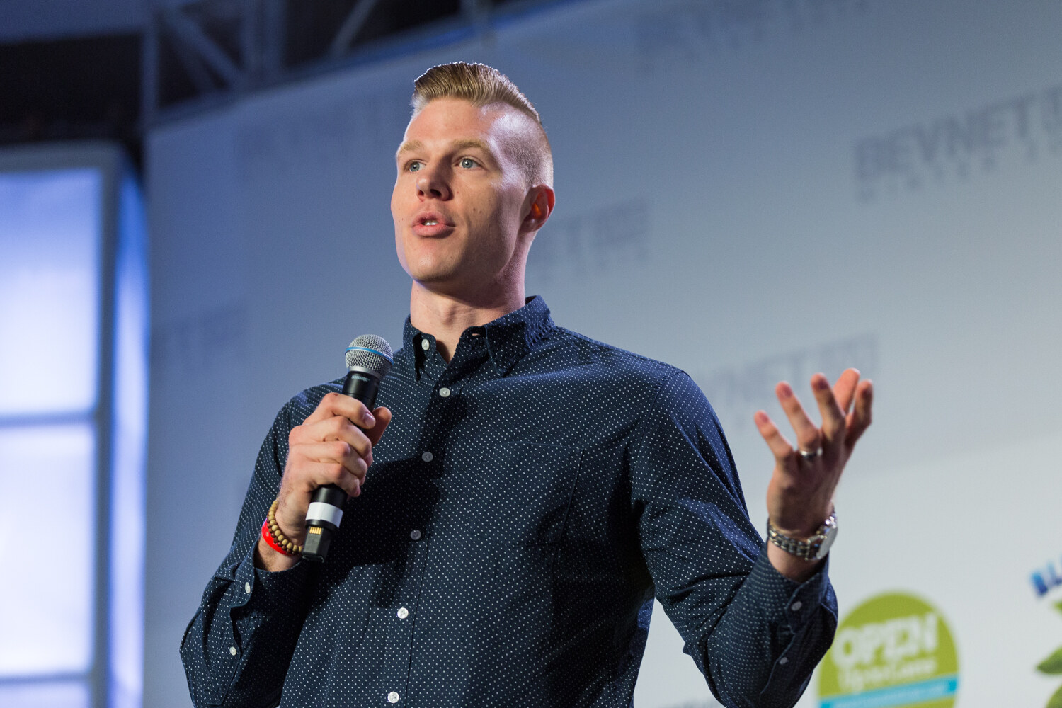 A man with a swept-back blond hairstyle in a navy polka-dot shirt holds a handheld microphone and raises his other hand in front of the sponsor wall.