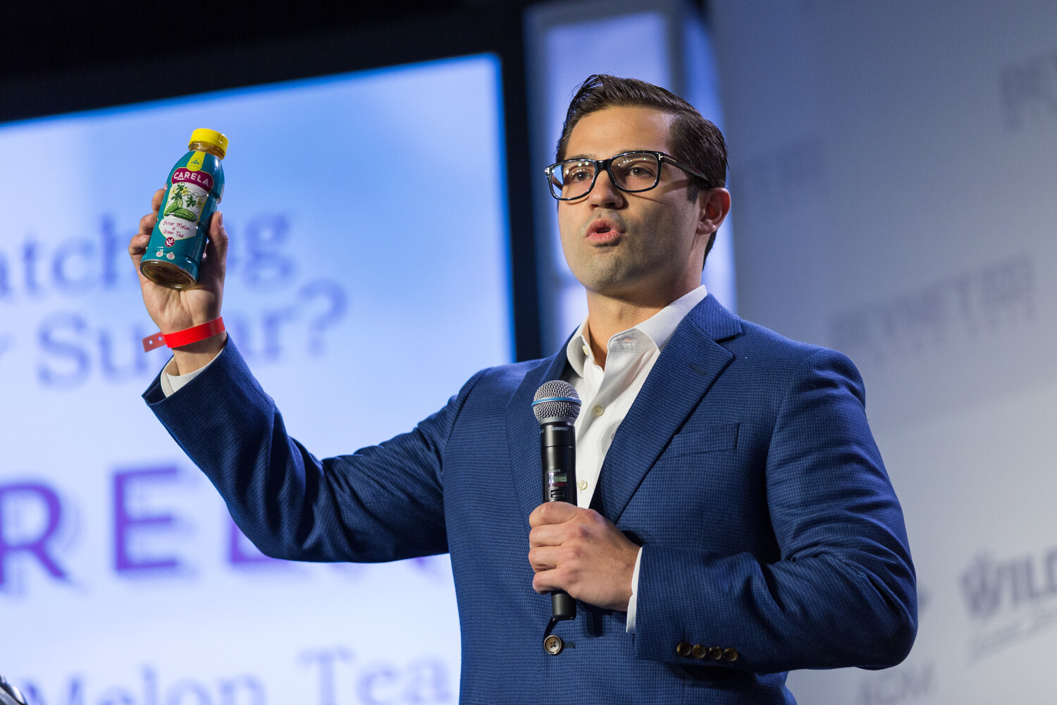 A man in glasses and a blue blazer holds a microphone in one hand and raises a teal Carela bottle with a yellow cap in the other, with a Watching Your Sugar and Melon Tea slide behind him.