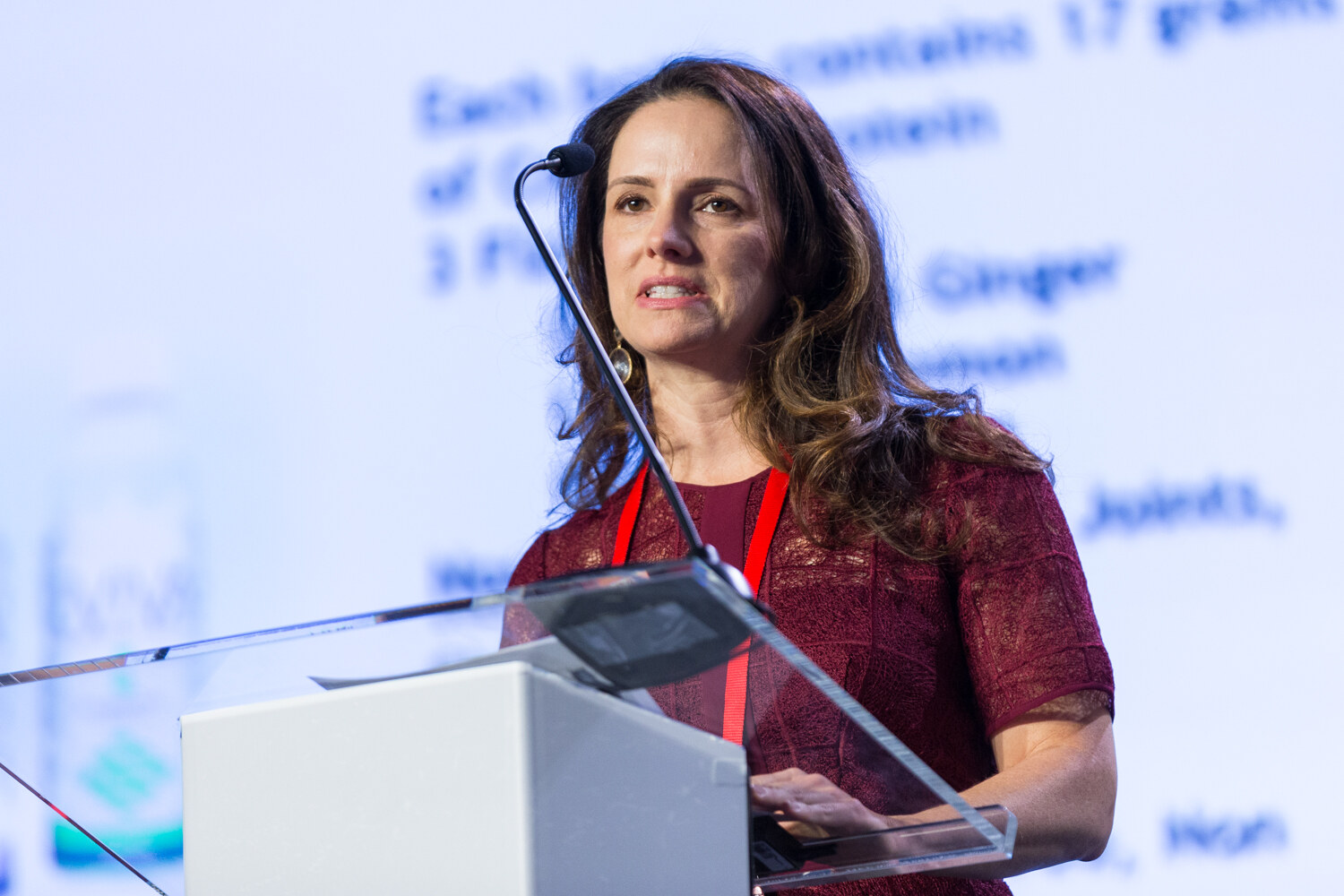 A woman with long wavy brown hair in a burgundy dress and red lanyard speaks at a clear lectern with a blurred slide mentioning 17 grams of protein, ginger and lemon behind her.