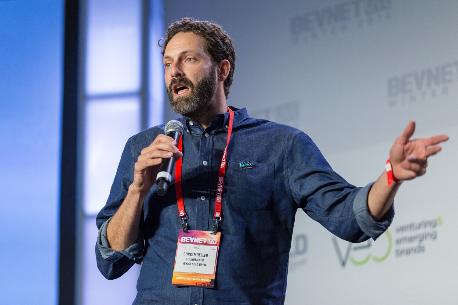 A bearded man in a denim shirt with a Venice pin and a BevNET Live badge reading Venice Cold Brew holds a microphone and points to his right.