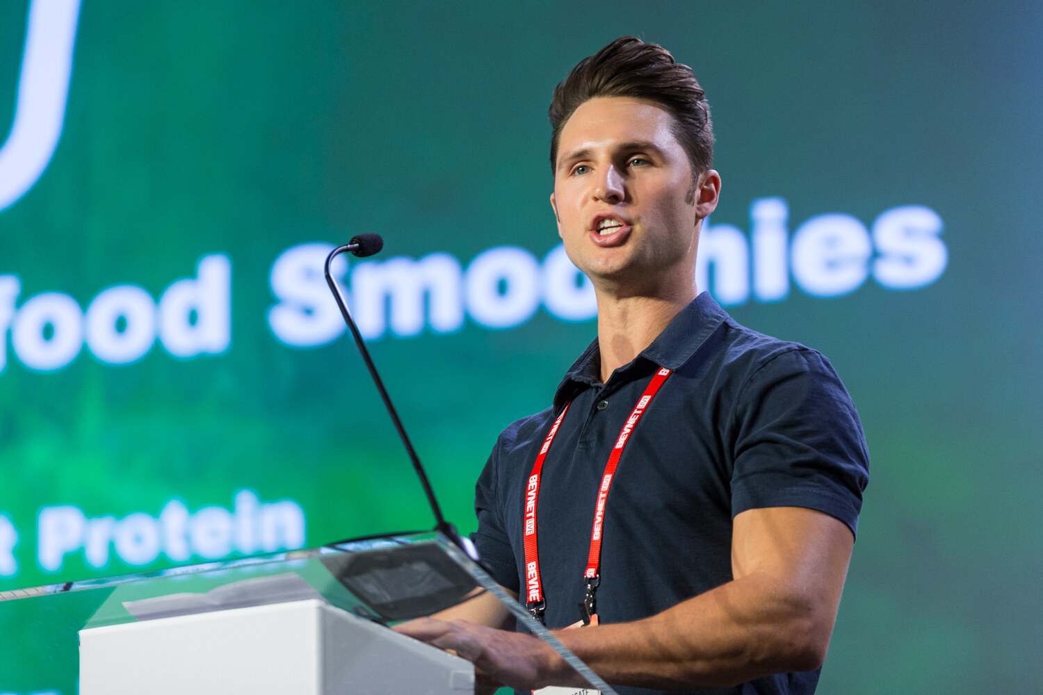 A young man in a navy polo and a red BevNET Live lanyard speaks at a clear lectern with a green screen reading Superfood Smoothies and Plant Protein behind him.